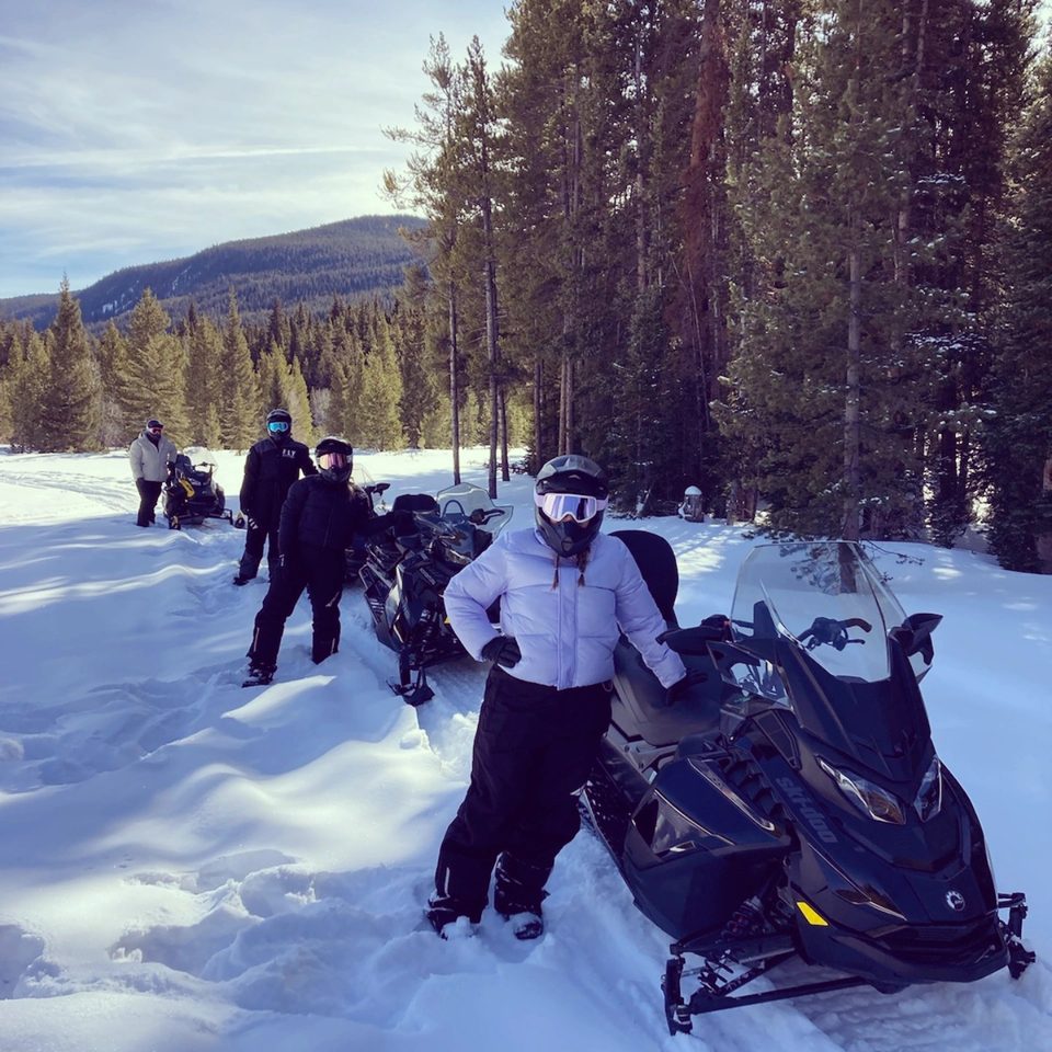 A guided group in helmets and goggles standing with black Ski-Doo touring snowmobiles in deep snow, pines and a forested ridge behind