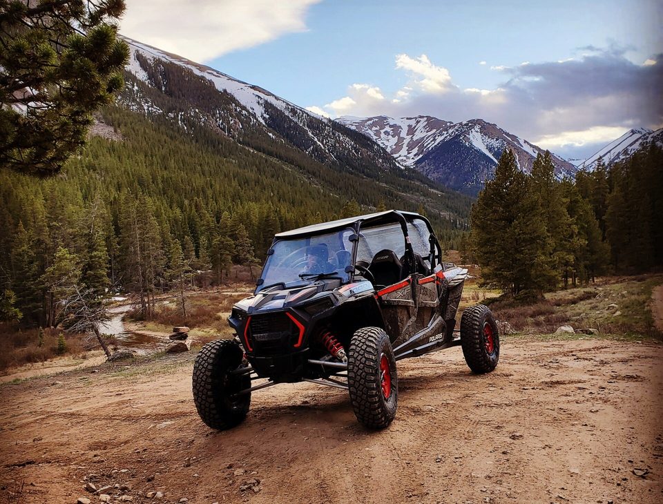 A black and red Polaris RZR XP 1000 parked on a dirt road beside a creek, snow-capped peaks behind