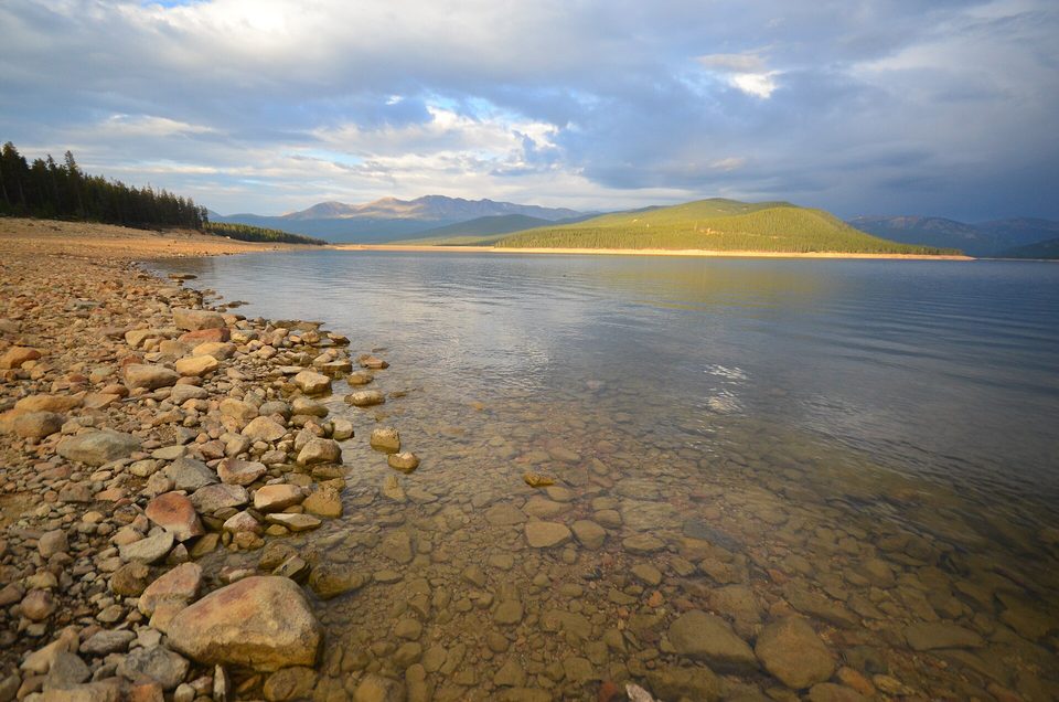 Rocky shoreline and calm water at Turquoise Lake with a forested peninsula lit by low sun